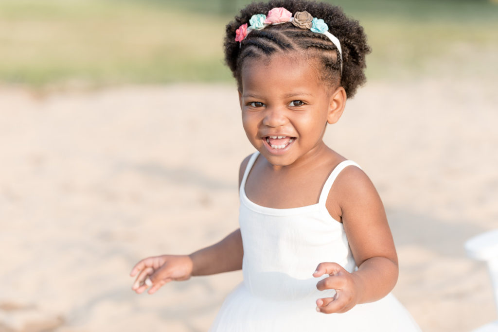 Tiny girl in white dress laughs during beach photo session.