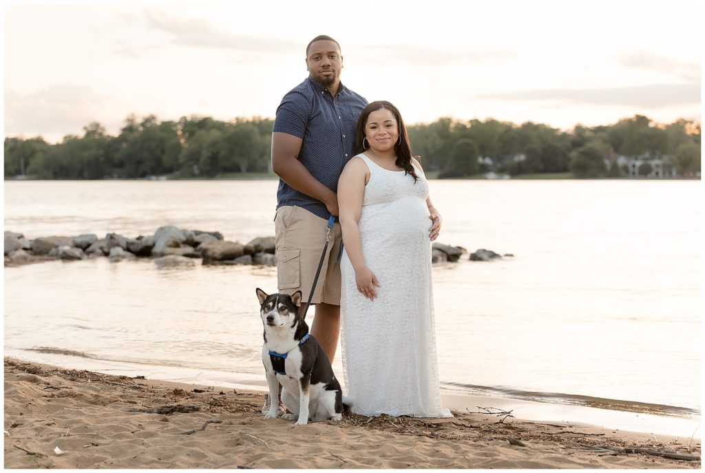 Husband, pregnant wife and family dog stand at water for photos