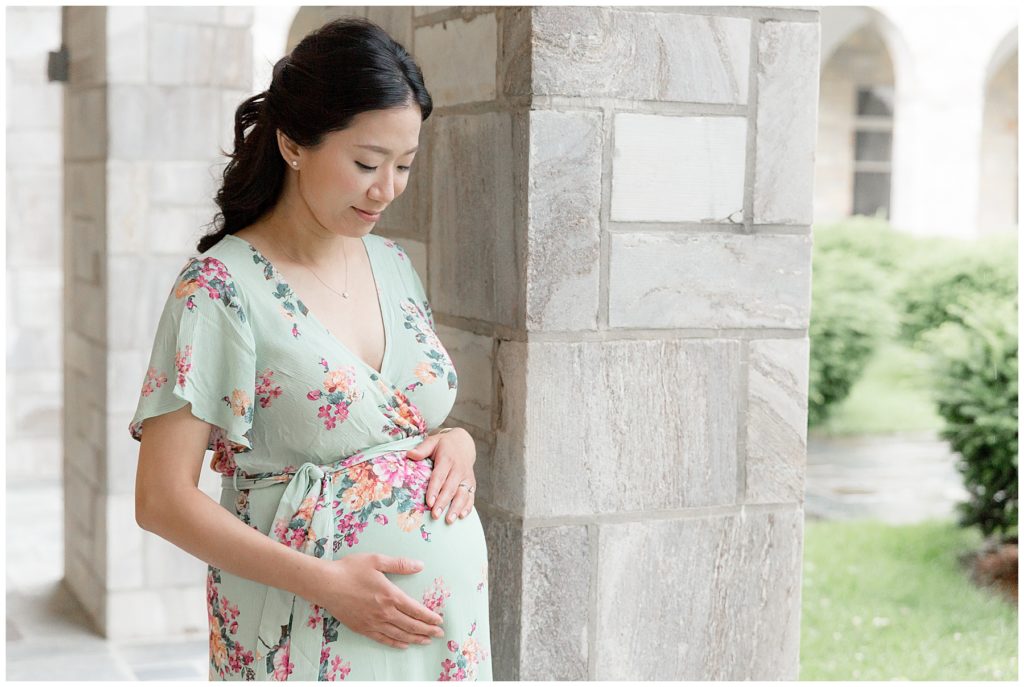 Beautiful pregnant momma in green flowered dress stands next white brick column
