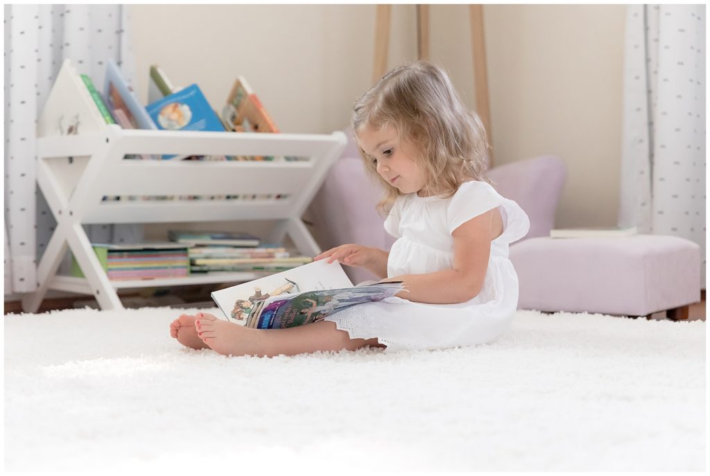 toddler sits on white rug and reads a book