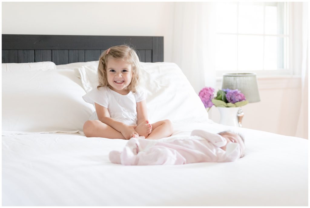 toddler sits on white master bed with newborn sister