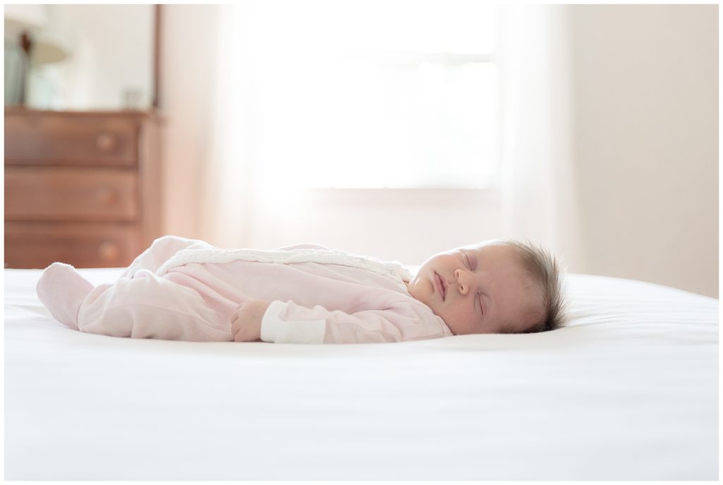 baby lays on white bedspread with large window providing backlight