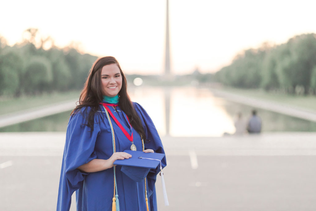Woman poses in blue cap and gown by reflecting pool and Washington Monument.
