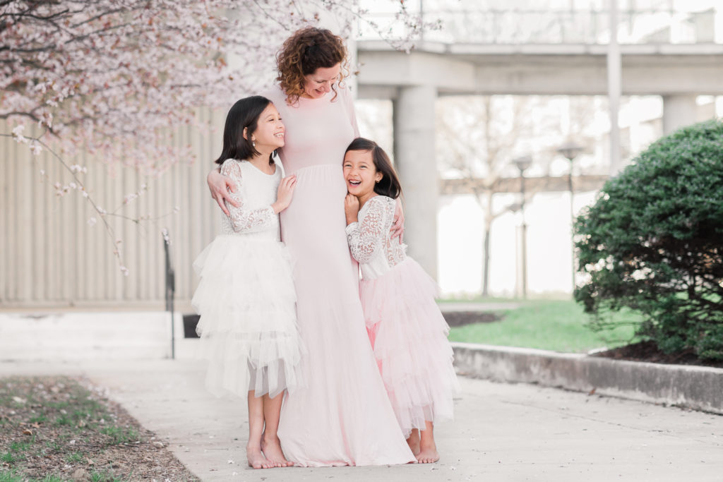 Mom and daughters in matching dresses smile at each other under the blooming cherry trees