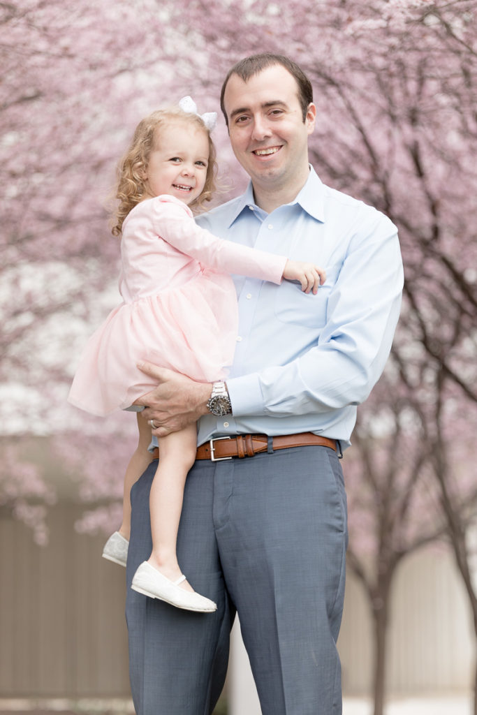 Dad holds toddler daughter under blooming cherry blossoms