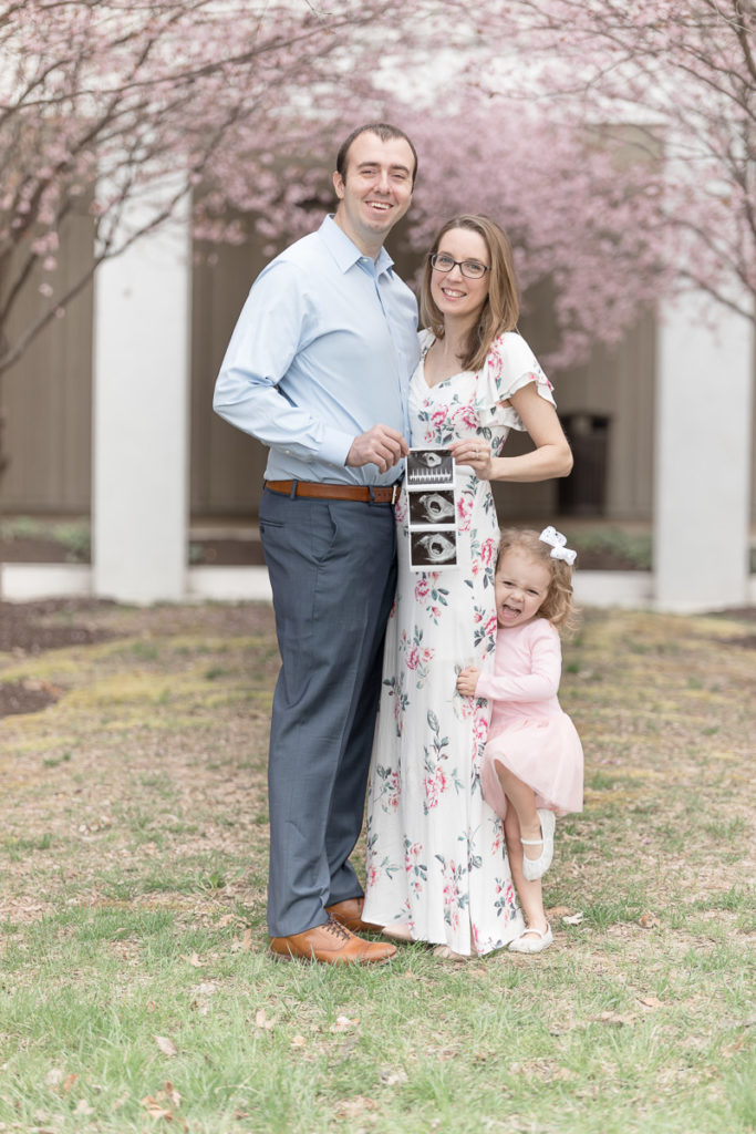 Family of three stands under spring blooms holding ultrasound photos to make their pregnancy announcement