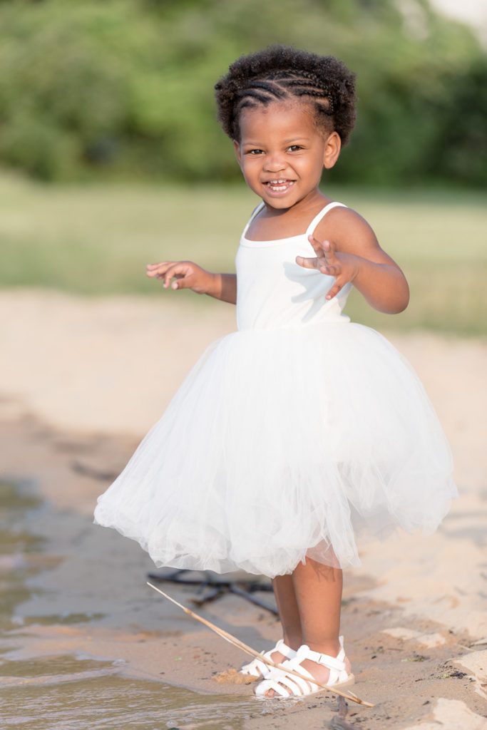 Toddler stands at the edge of the water in white dress and white sandals.