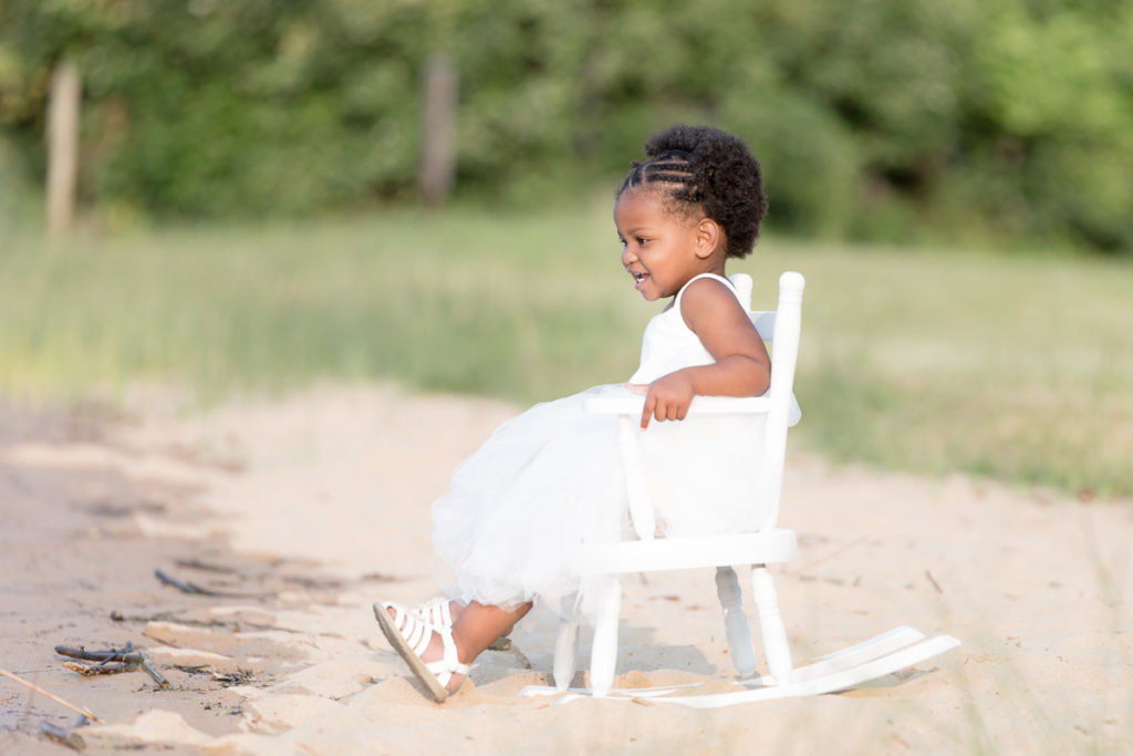 Little girl sits in white rocking chair in morning photo session