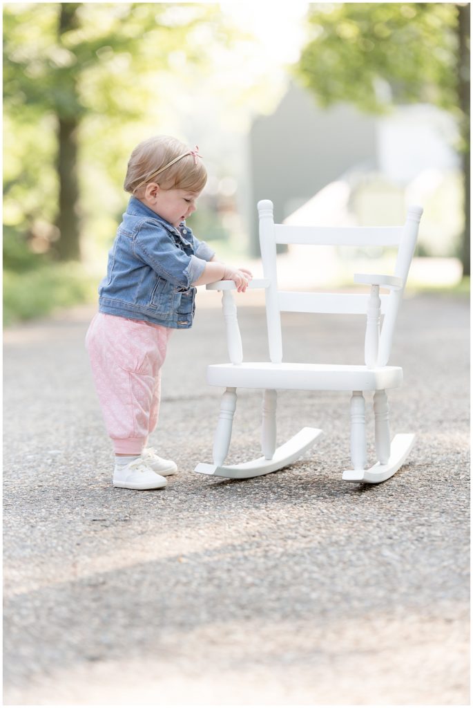 One year old girl stands next to small white rocking chair