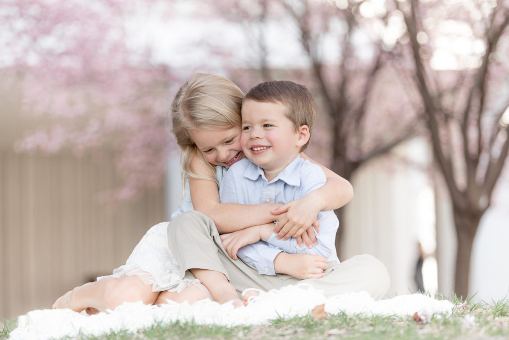 big sis hugs little brother while surrounded by cherry blossoms