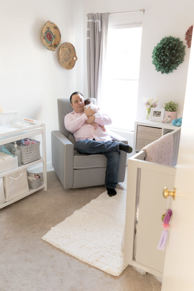 Dad sits in nursery rocker and pats his baby's back during their lifestyle newborn photos