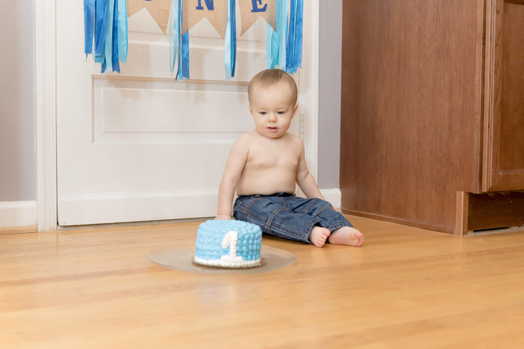 One year old boy sits on floor looking at smash cake
