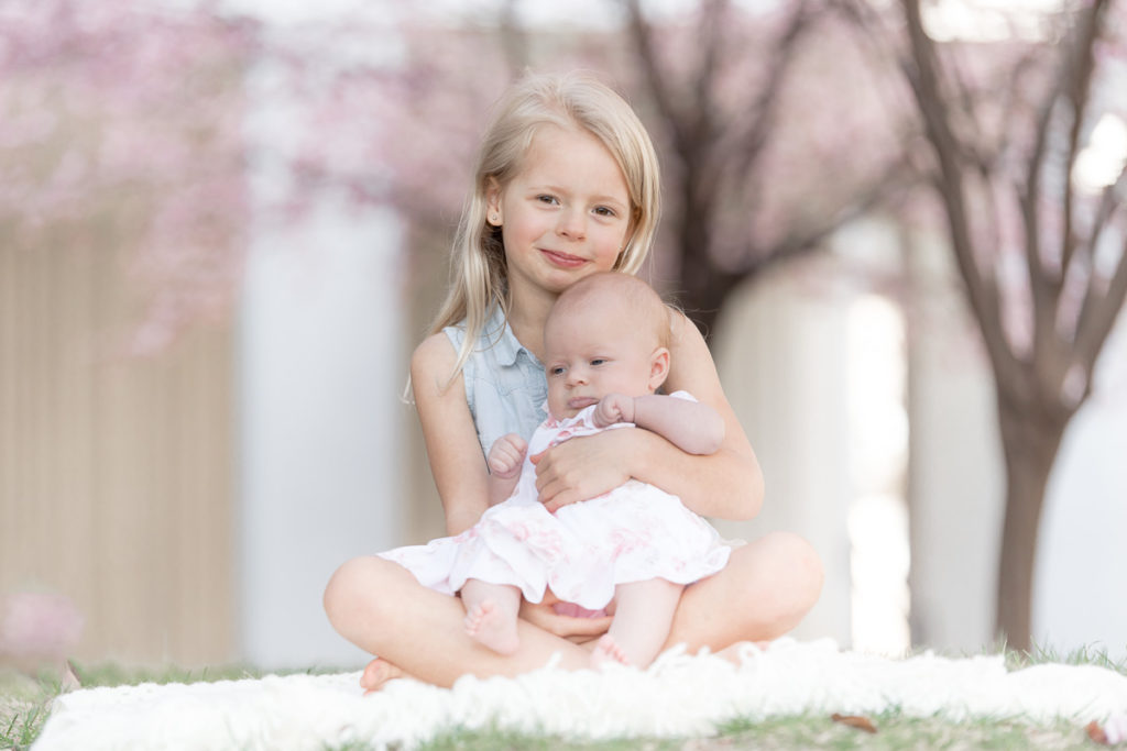 big sister snuggles her baby sister in the shade of the cherry blossom trees