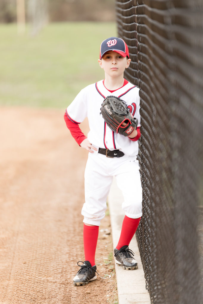 boy dressed in red/white baseball uniform leans against fence