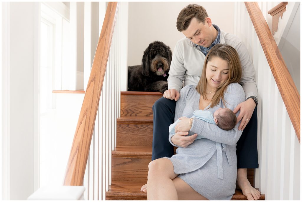 parents and dog sit in light-filled stairwell with new baby