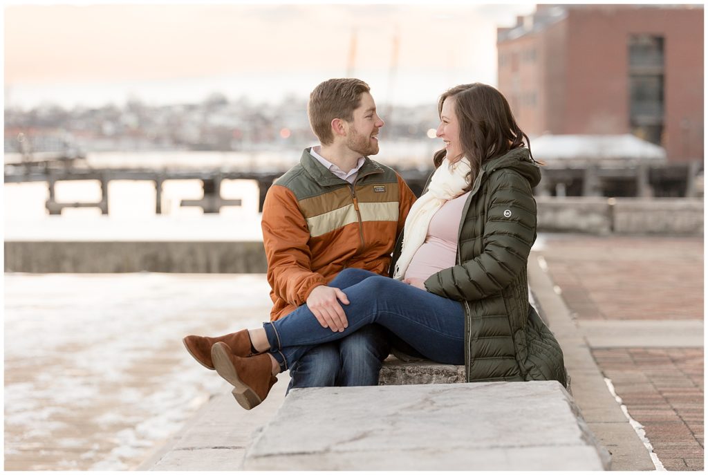 pregnant couple sits on stone bench in front of Baltimore sunset
