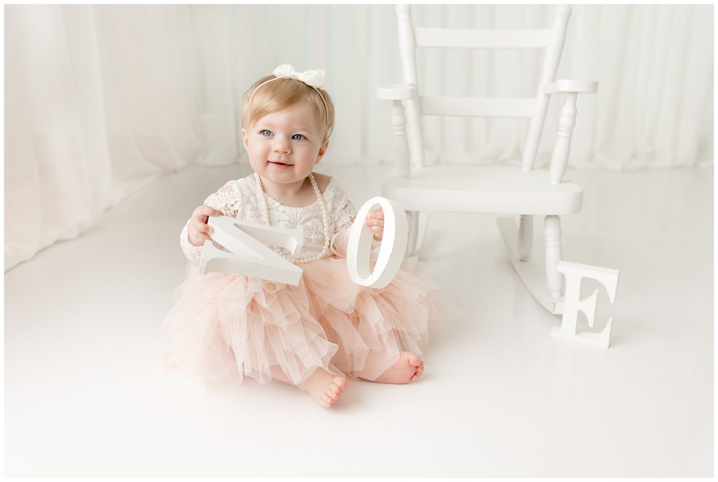blond baby in puffy pink dress holds letter blocks spelling ONE