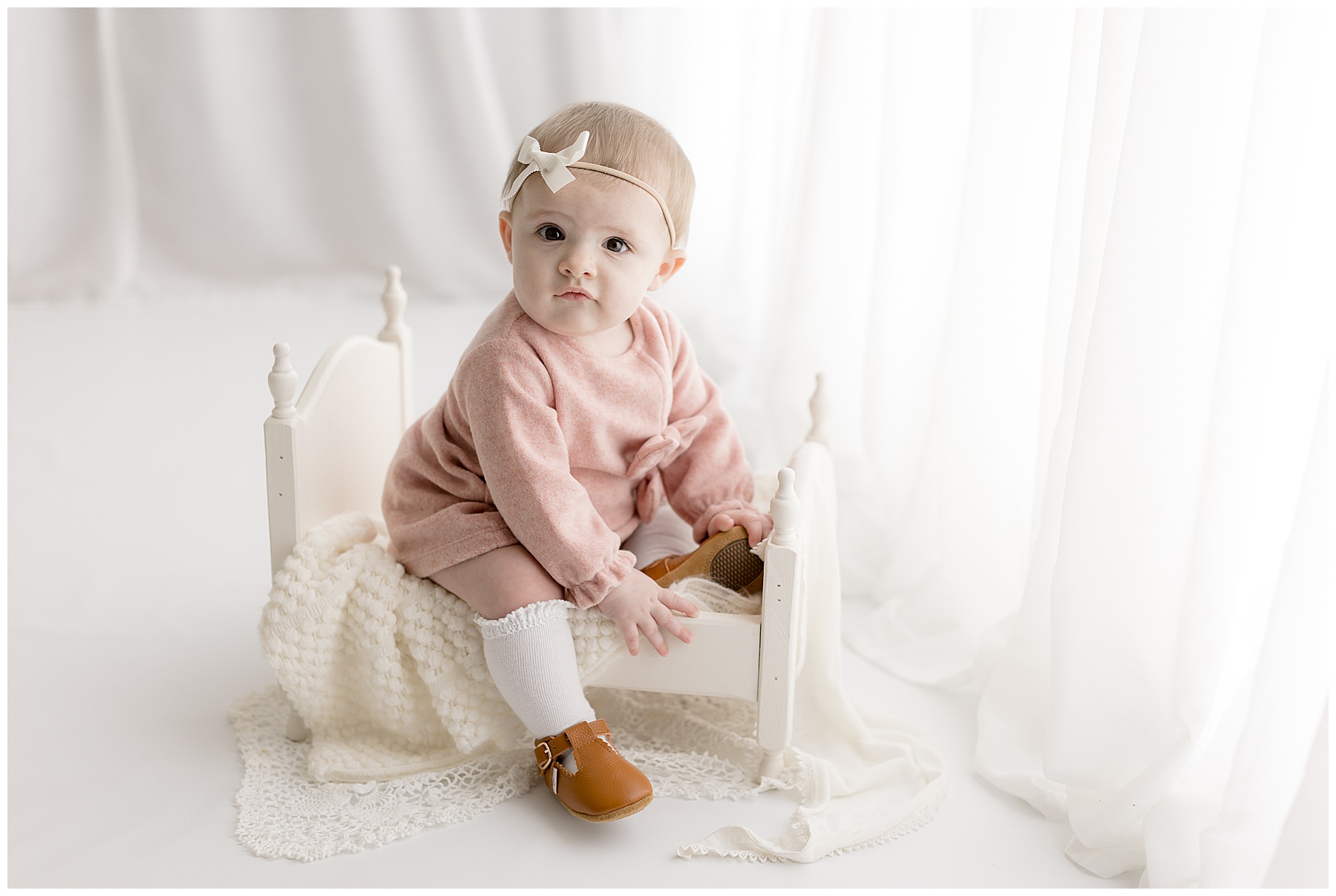 A dainty baby girl in pink dress, knee socks, and brown leather shoes sits on a white wooden bed prop in front of a window with sheer white curtains.