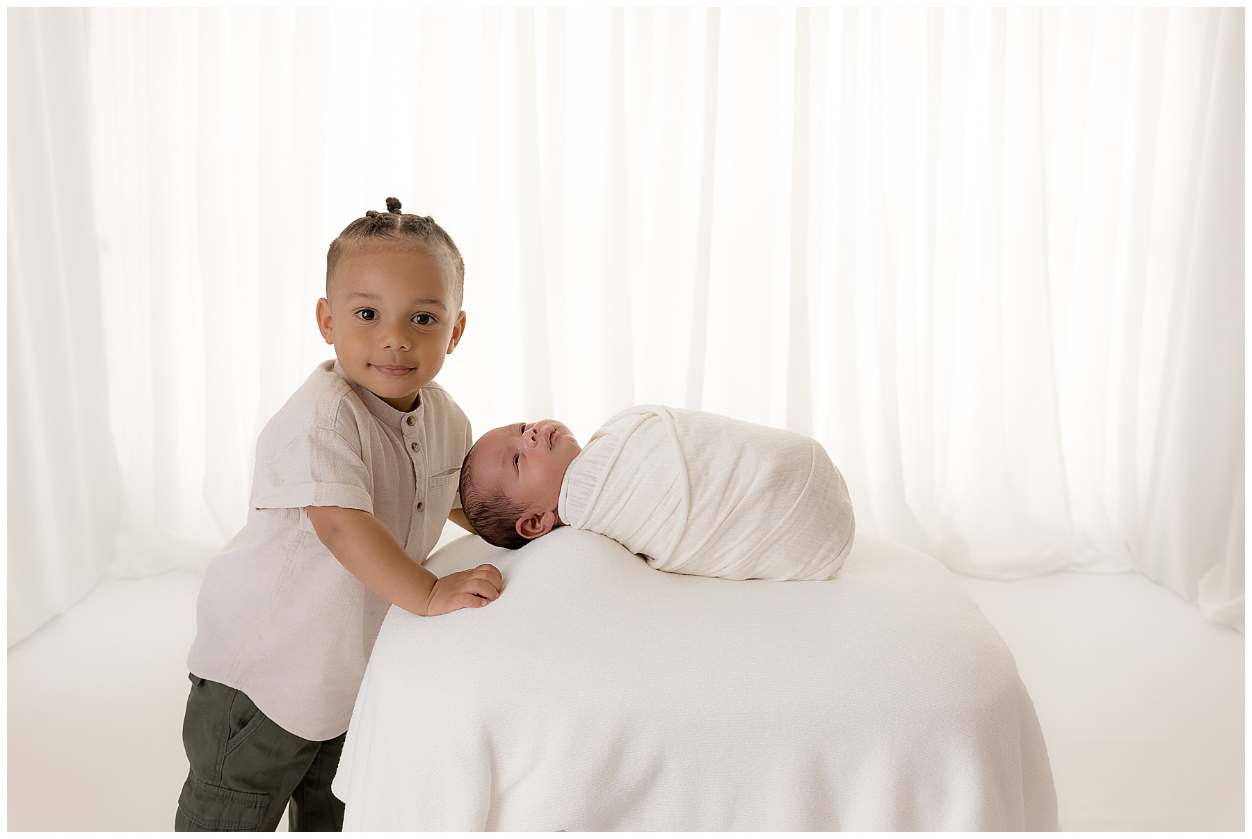 A toddler and a newborn photographed together in a white room.