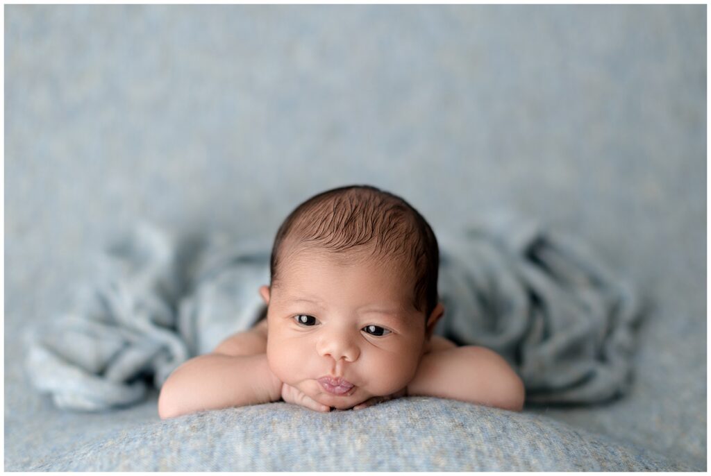 An awake baby lies on a blue backdrop with his sweet little lips pursed. The power of photos means that his momma will remember this moment always.