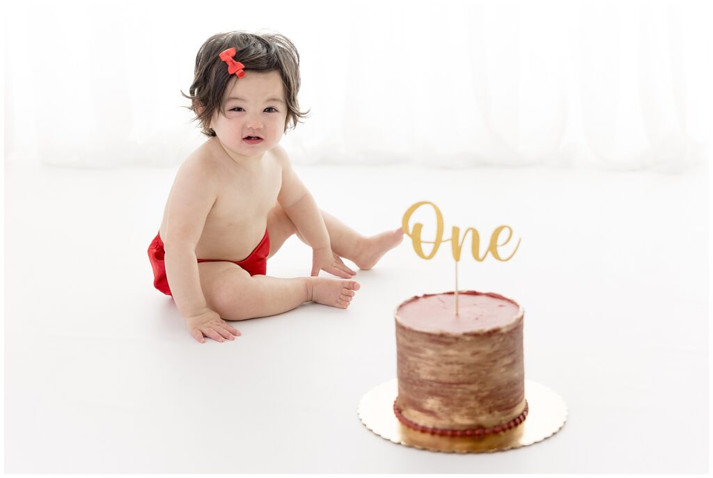 Silly baby makes a face at the camera while sitting in front of her red and gold smash cake.