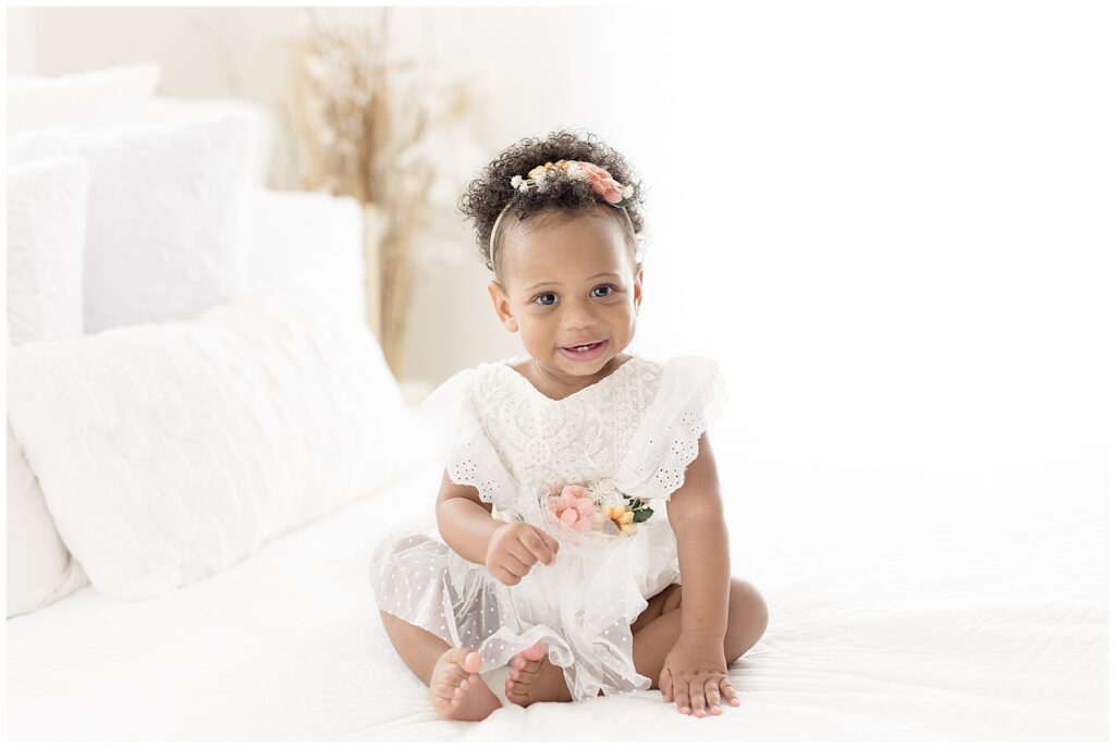 baby girl in white dress with flower details sits on white bed