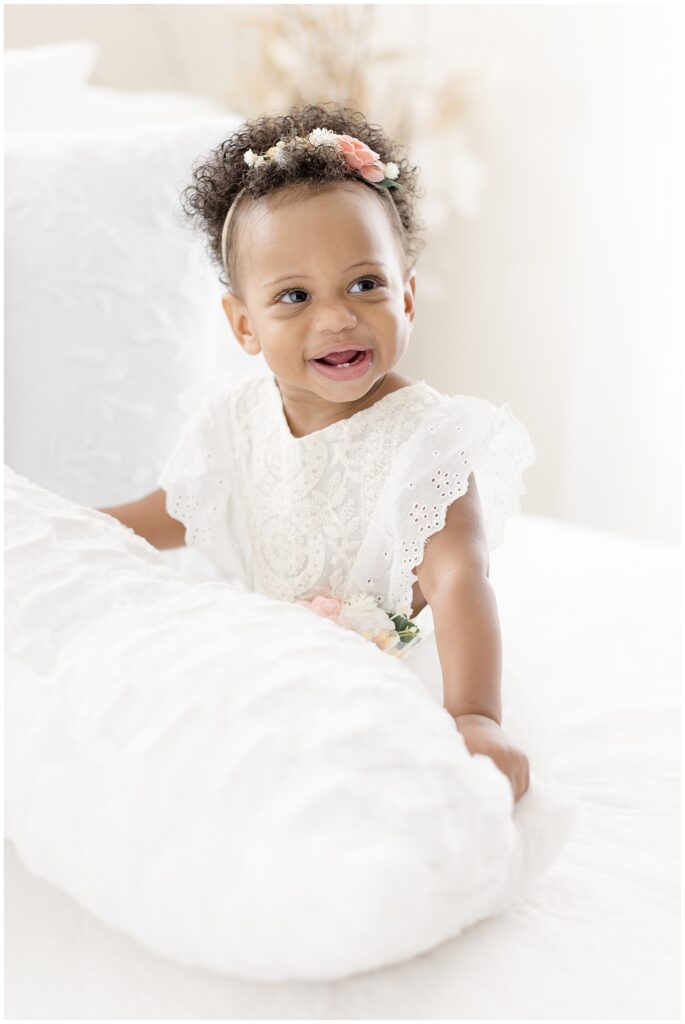 cute one year old girl sits on a white bed and plays with a pillow while laughing
