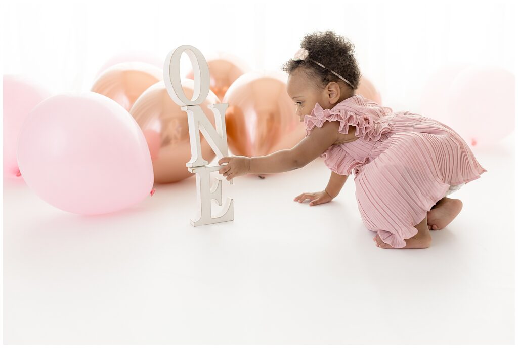 baby girl, surrounded by balloons, reaches for wooden letters that spell out ONE